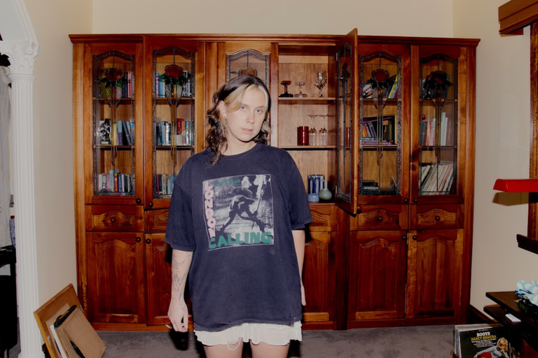 Musician Grace Robinson wearing a London Calling t-shirt standing in front of a wooden display cabinet.